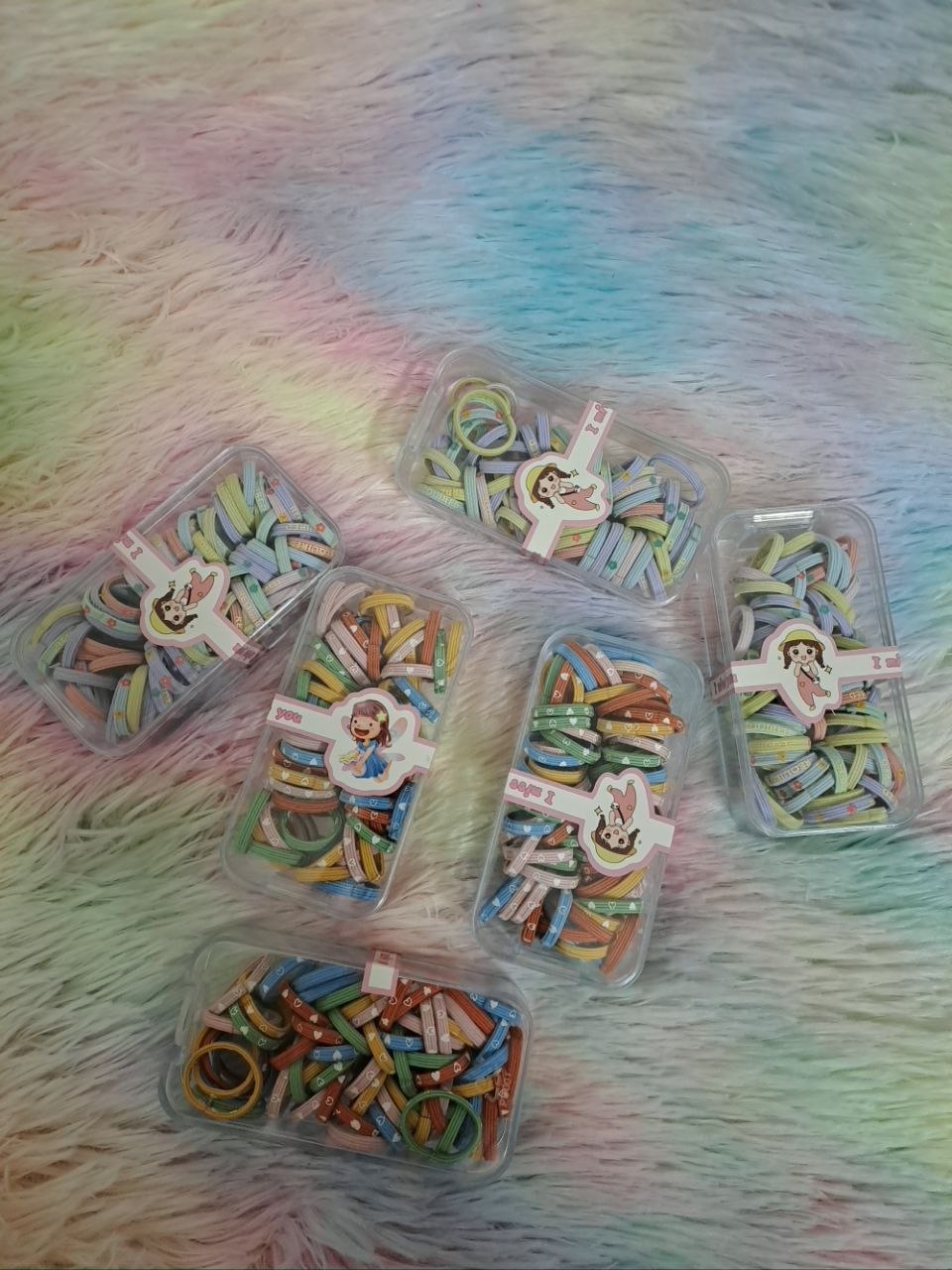 Clear storage containers filled with colorful rainbow thin elastic hair ties in various solid and multicolor combinations for girls