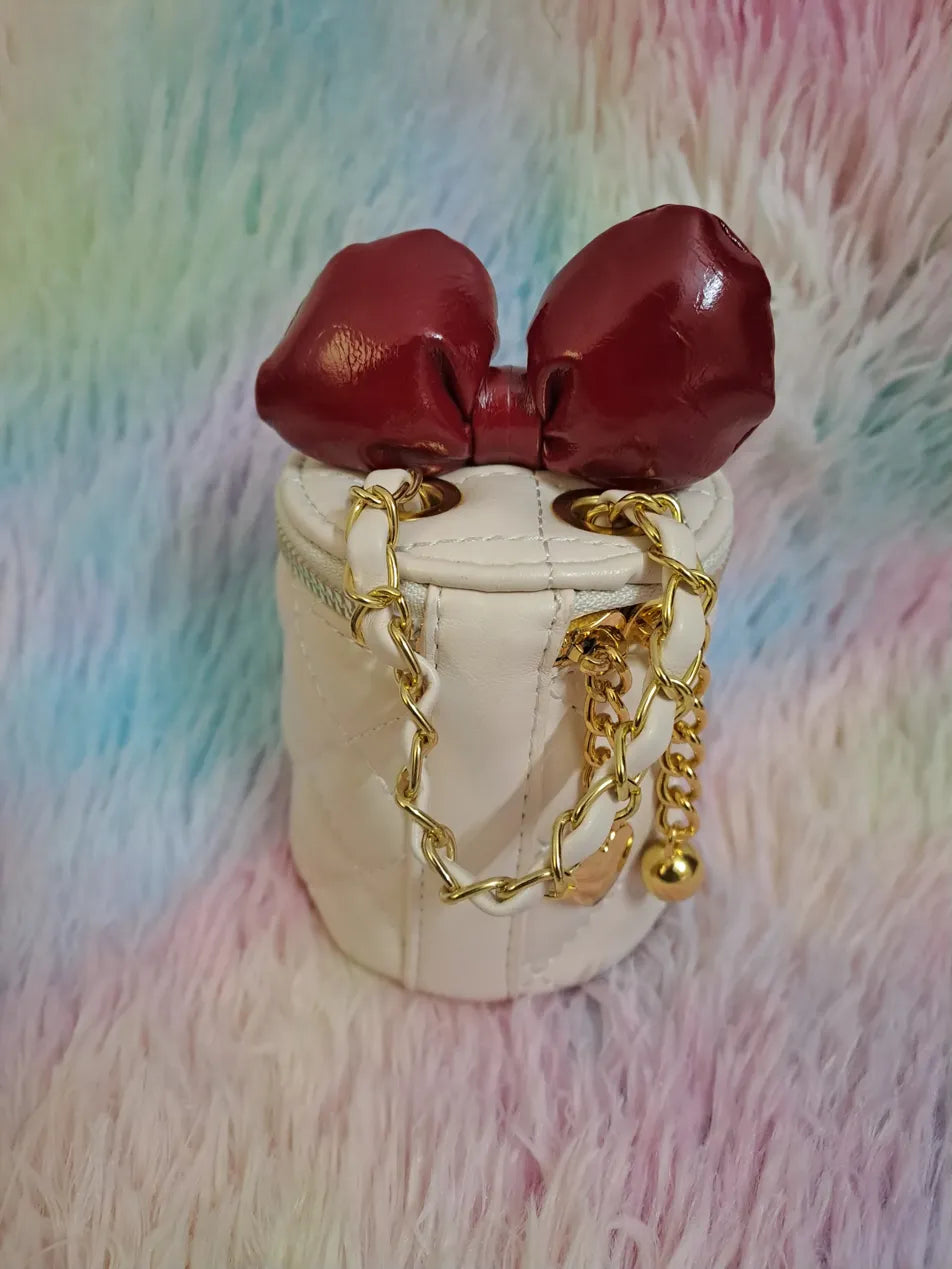 Cream quilted cylinder bag with rich burgundy bow closure woven through dual gold chain handles for girls
