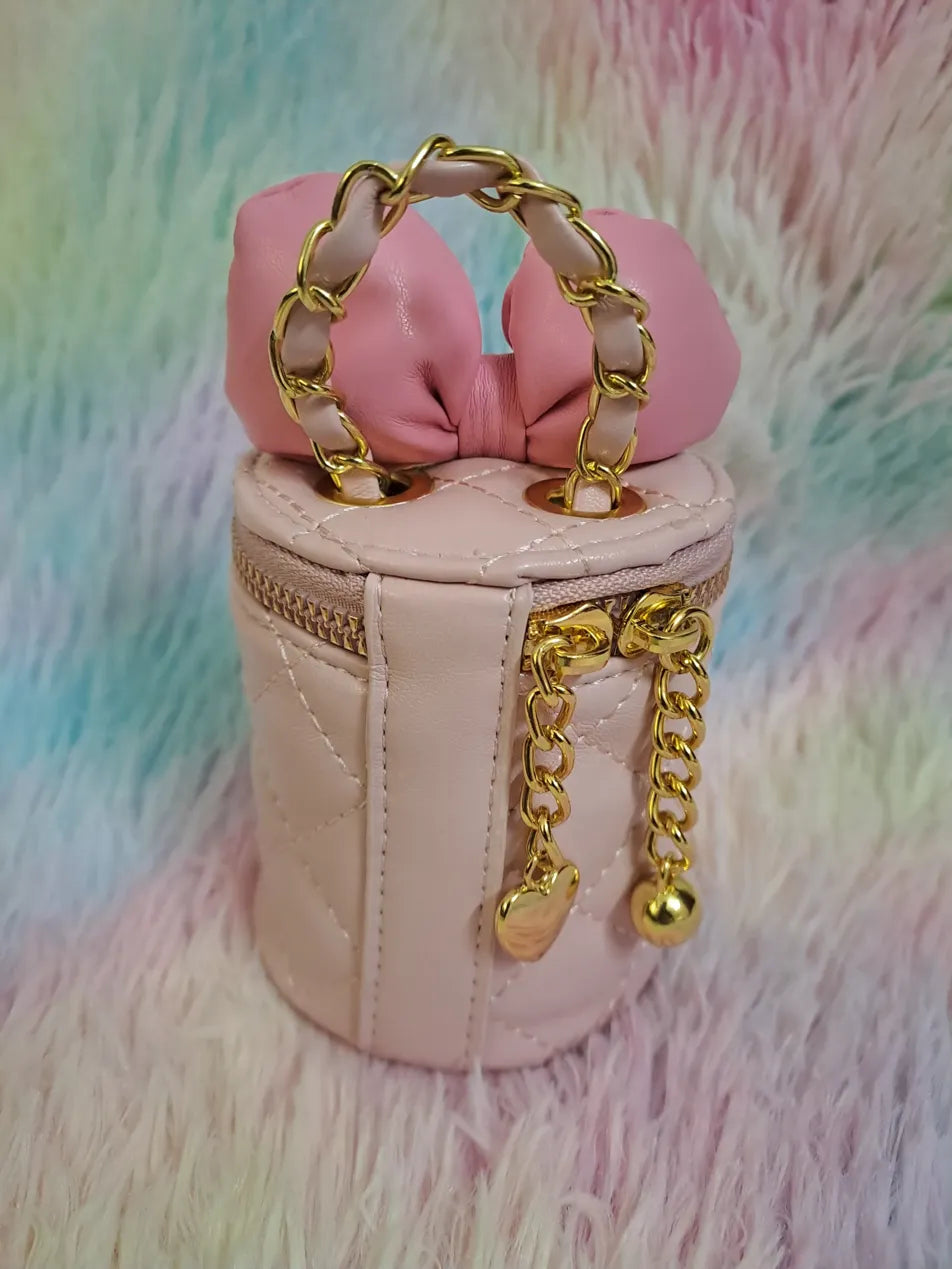 Beige quilted cylinder bag with pink bow closure woven through dual gold chain handles for girls