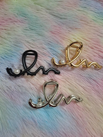 Scripted calligraphy wave pearl hair claw clips in matte black, warm gold, and polished silver finishes, with freshwater pearl accent, spring-loaded jaw mechanism, elegant sculptural metal hair accessory
