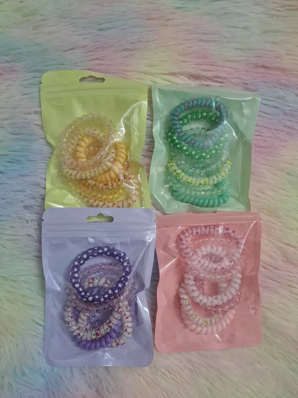 Spiral phone cord coil hair ties in four pastel packs: yellow with seasonal prints, green-teal with floral polka dot prints, purple with snowman holiday prints, and pink with romantic patterns, multiple coils per pack in zip resealable bags