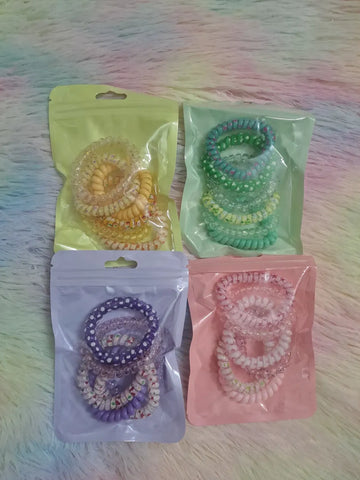 Spiral phone cord coil hair ties in four pastel packs: yellow with seasonal prints, green-teal with floral polka dot prints, purple with snowman holiday prints, and pink with romantic patterns, multiple coils per pack in zip resealable bags