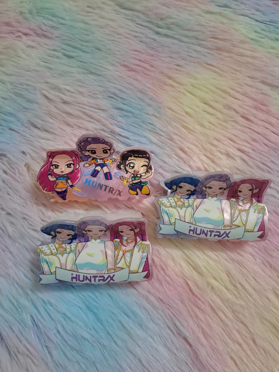 HUNTRX acrylic character hair clip badge set of 3 designs featuring chibi action-pose characters and elegant full-group portrait designs with high-quality printed artwork under clear protective acrylic, fan merchandise hair accessories